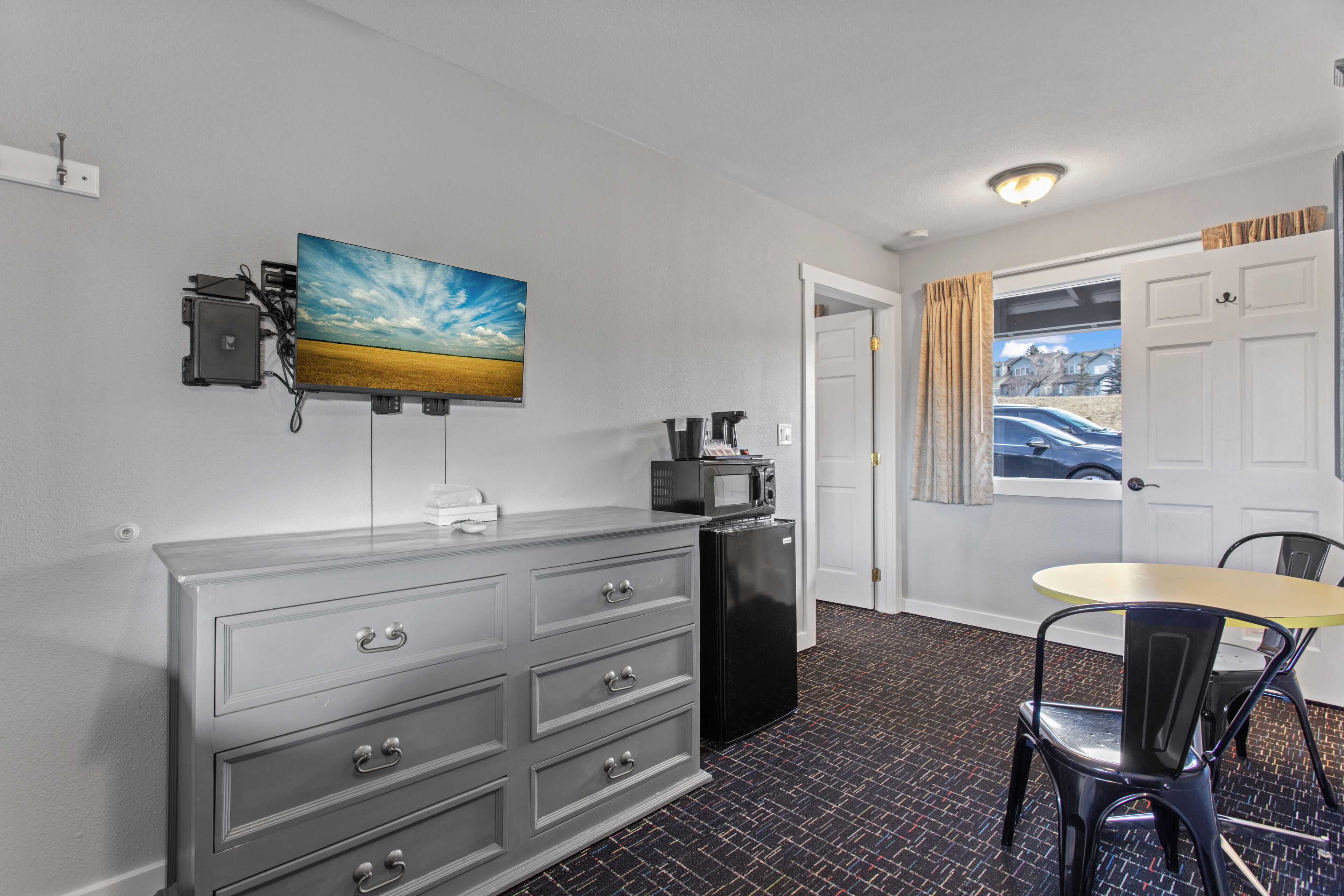 A compact hotel room with a kitchenette, a dining area with a table and chairs, a television mounted on the wall, and a door leading to an outdoor area.