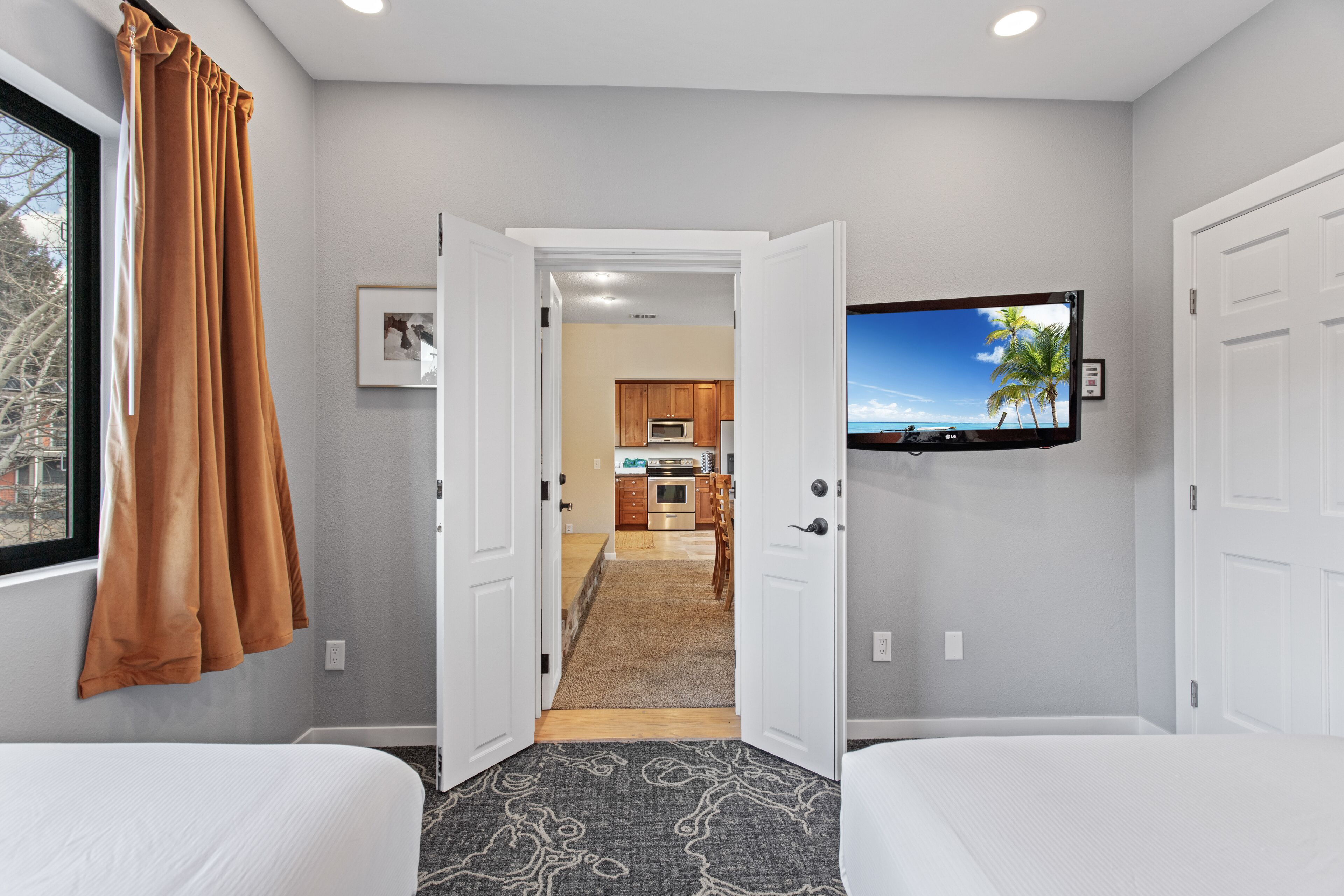 A hotel room with two beds, a TV mounted on the wall, a kitchen in the background, and a view of palm trees through the window.