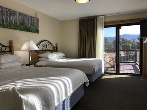 A hotel room with two beds, a painting on the wall, a lamp, and a view of mountains through the window. - Coyote Mountain Lodge (Estes Park)
