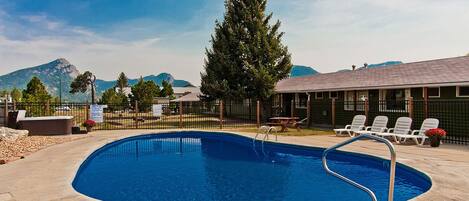 A swimming pool with a metal ladder, surrounded by a fence, with lounge chairs and a building in the background.