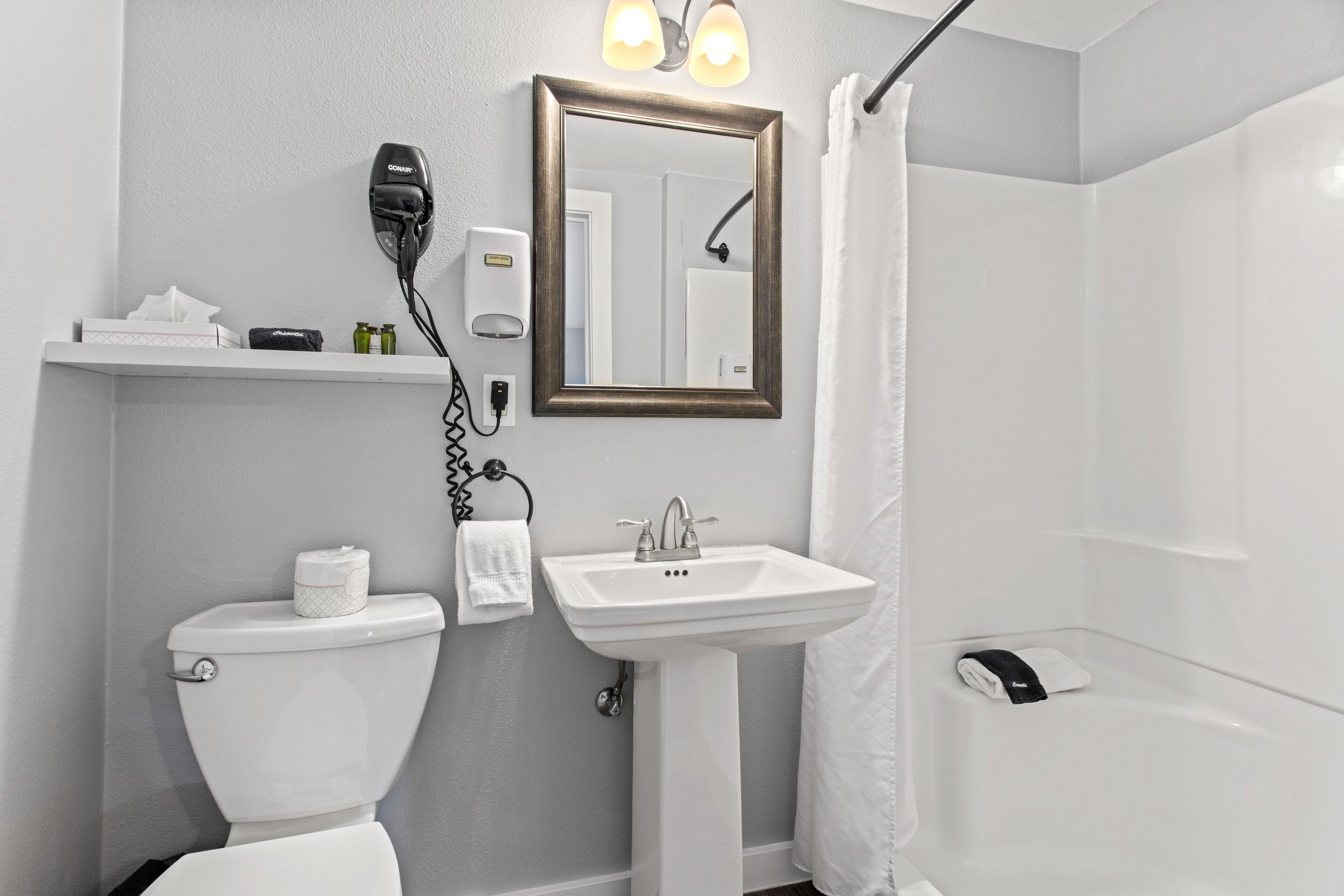 A bathroom with a toilet, sink, and shower. There is a hairdryer and a soap dispenser on the wall.