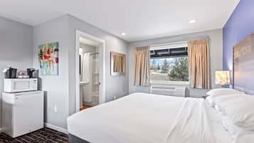 A hotel room with a bed, a microwave, a small refrigerator, a window with curtains, and a view of trees.