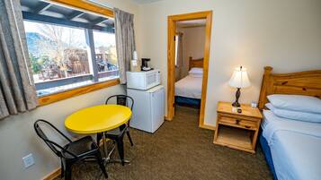 Une petite chambre d’hôtel avec un lit simple, une table de chevet avec une lampe, un coin cuisine compact, une petite table avec une chaise et une fenêtre avec des rideaux.