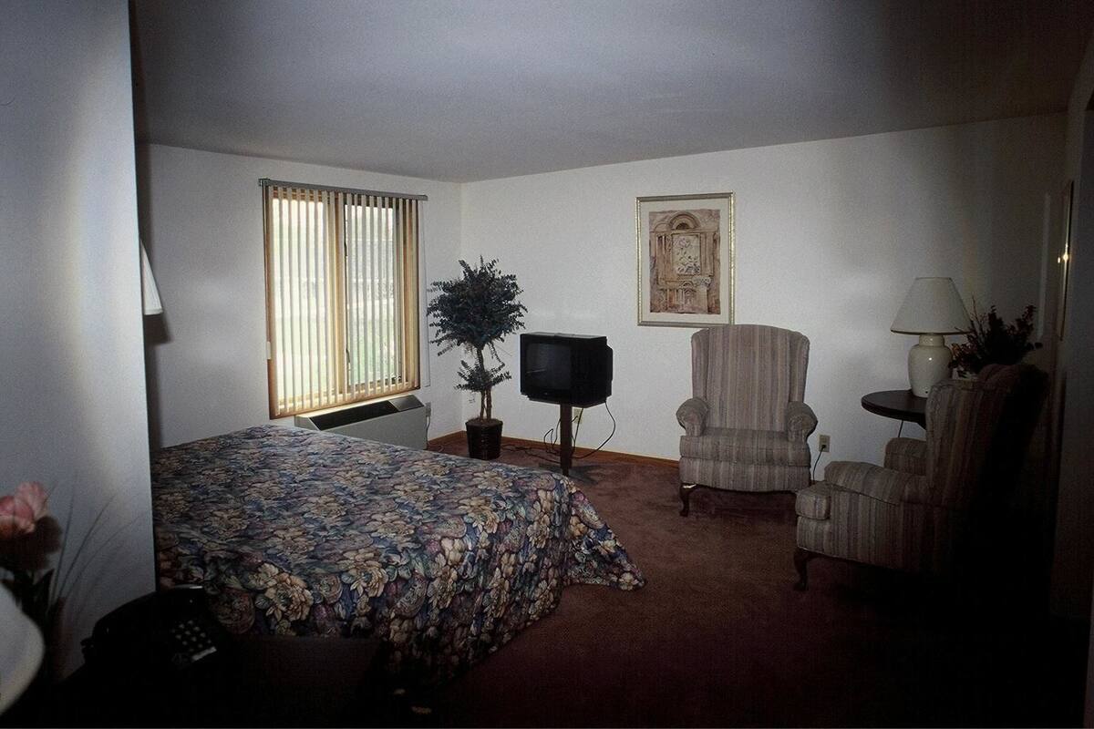 Una camera d'albergo con un letto, una televisione, una sedia, una lampada e una finestra con persiane.