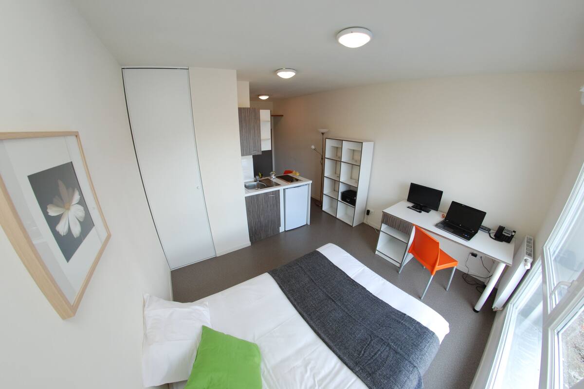 A modern hotel room with a bed, a desk with a computer, a chair, and a kitchenette.