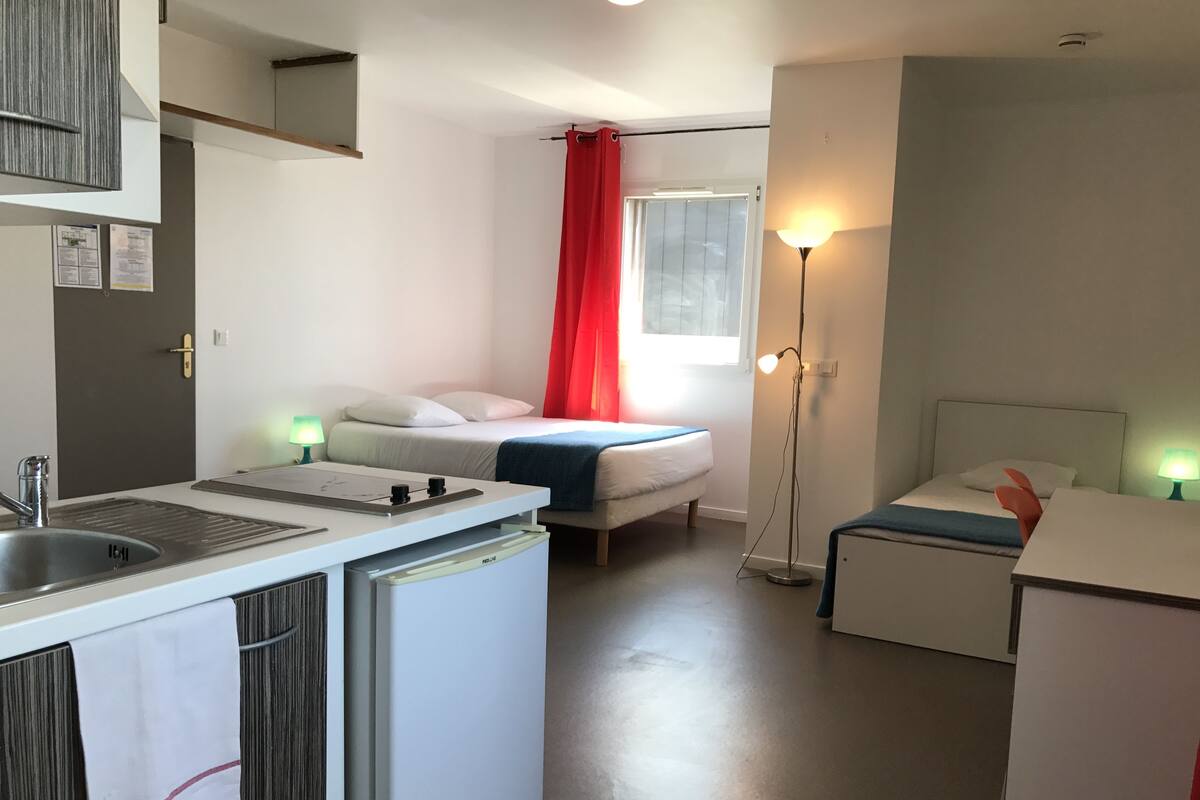 A compact hotel room with a kitchenette, a bed with a red curtain, a bedside table with a lamp, and a small desk with a lamp.