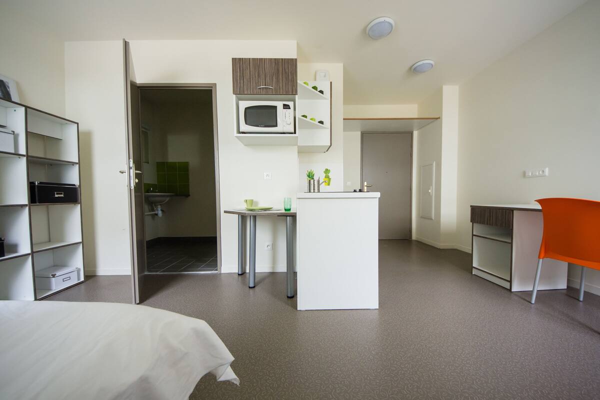 A compact hotel room with a bed, a small kitchenette, a desk, and a chair.