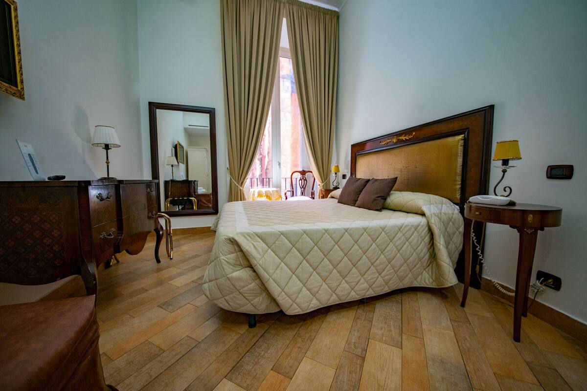 A hotel room with a large bed, wooden flooring, a dresser, a mirror, and a window with curtains.