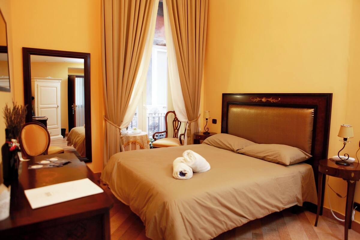 A hotel room with a bed, a desk with a mirror, a chair, and a window with curtains.