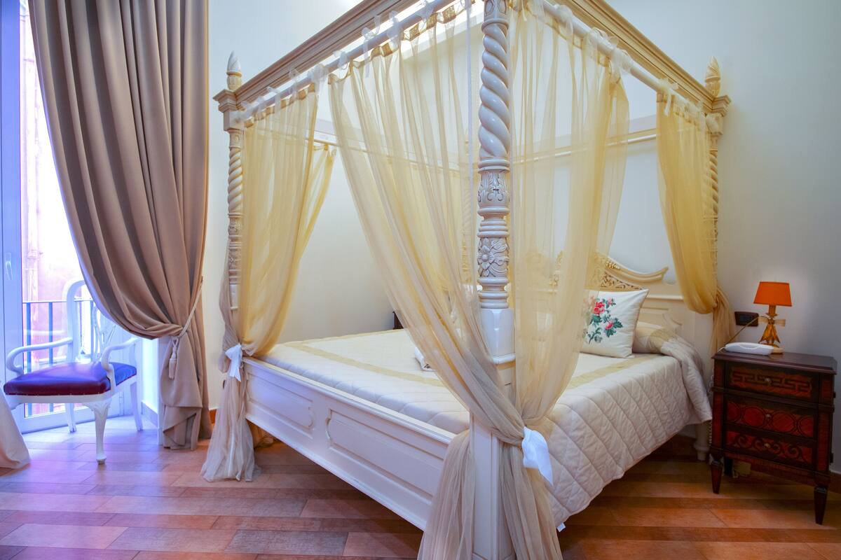 A four-poster bed with sheer curtains, a bedside table with a lamp, and a chair.