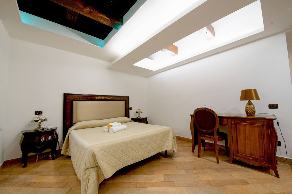 A bedroom with a bed, a desk, and a chair. There is a skylight above the bed.