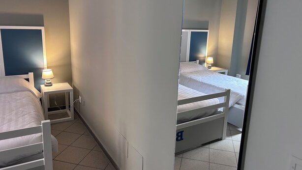 Una habitación de hotel con dos camas individuales, cada una con una mesita de noche y una lámpara. El piso es de baldosas y las paredes son blancas.