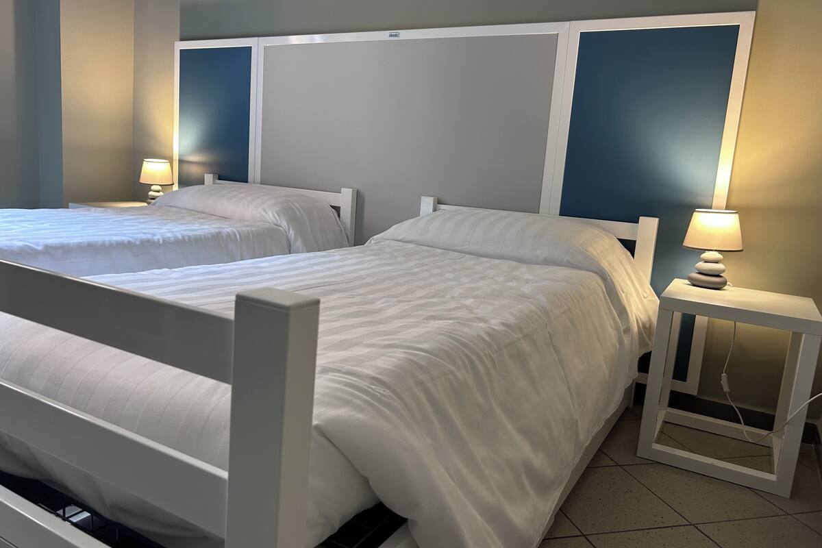 Habitación de hotel con dos camas, cada una con cabecero blanco y mesita de noche con lámpara.