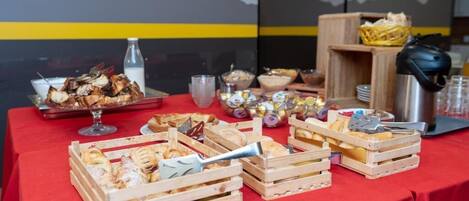 Uma mesa de buffet com diversos alimentos e utensílios, incluindo doces, pão e acompanhamentos, sobre uma toalha de mesa vermelha.