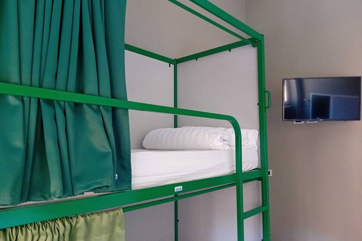 Una cama individual con barandillas de metal verde, un colchón blanco y una cortina verde. Un televisor está montado en la pared a la derecha.