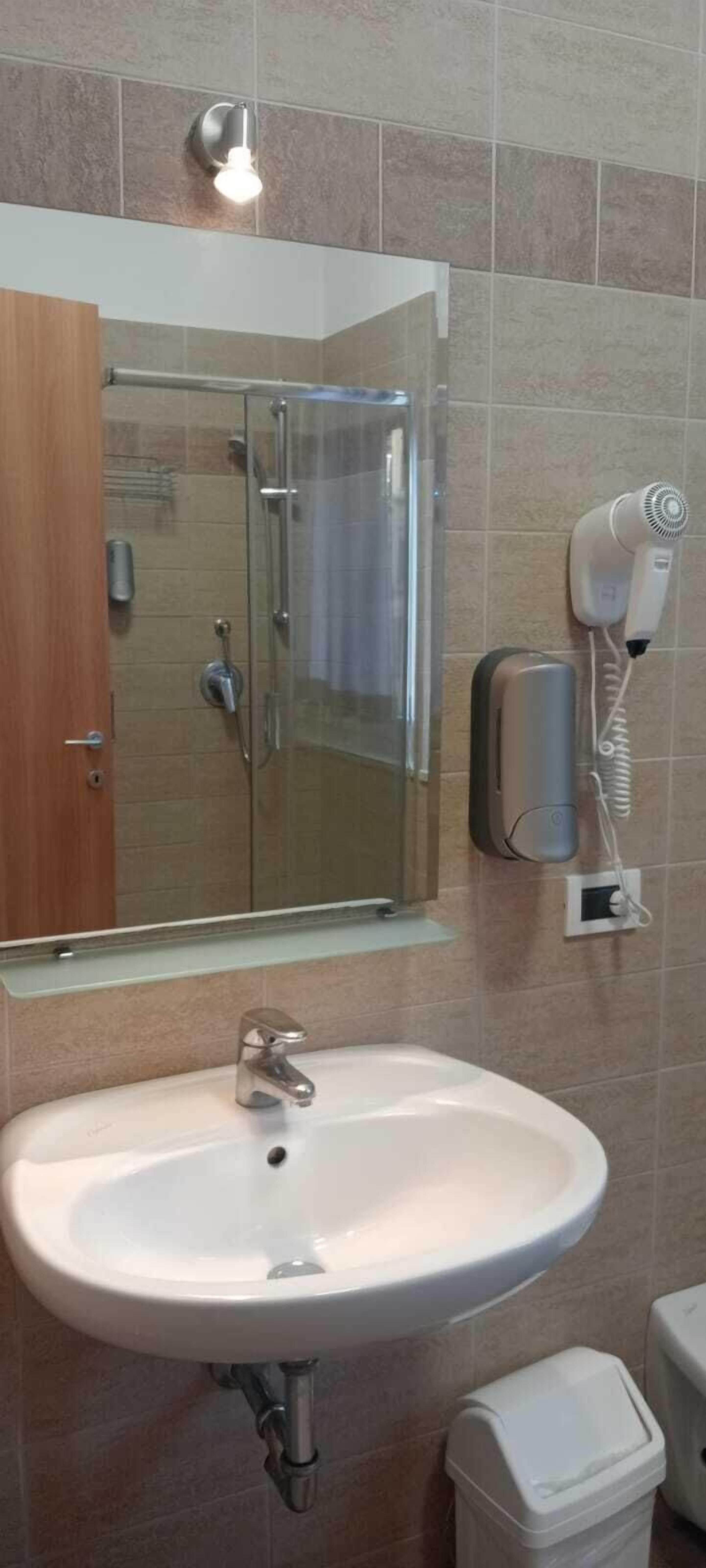 A bathroom with a glass shower enclosure, a white sink with a mirror above it, and a hairdryer mounted on the wall.