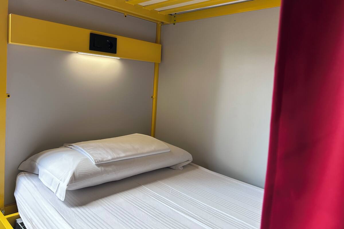 Una cama individual con cabecera, un estante pequeño arriba y una cortina roja parcialmente visible.