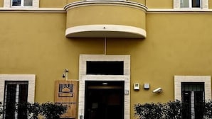 Un edificio con fachada amarilla, un balcón con ventanal circular y banderas de Italia, la Unión Europea y otro país.