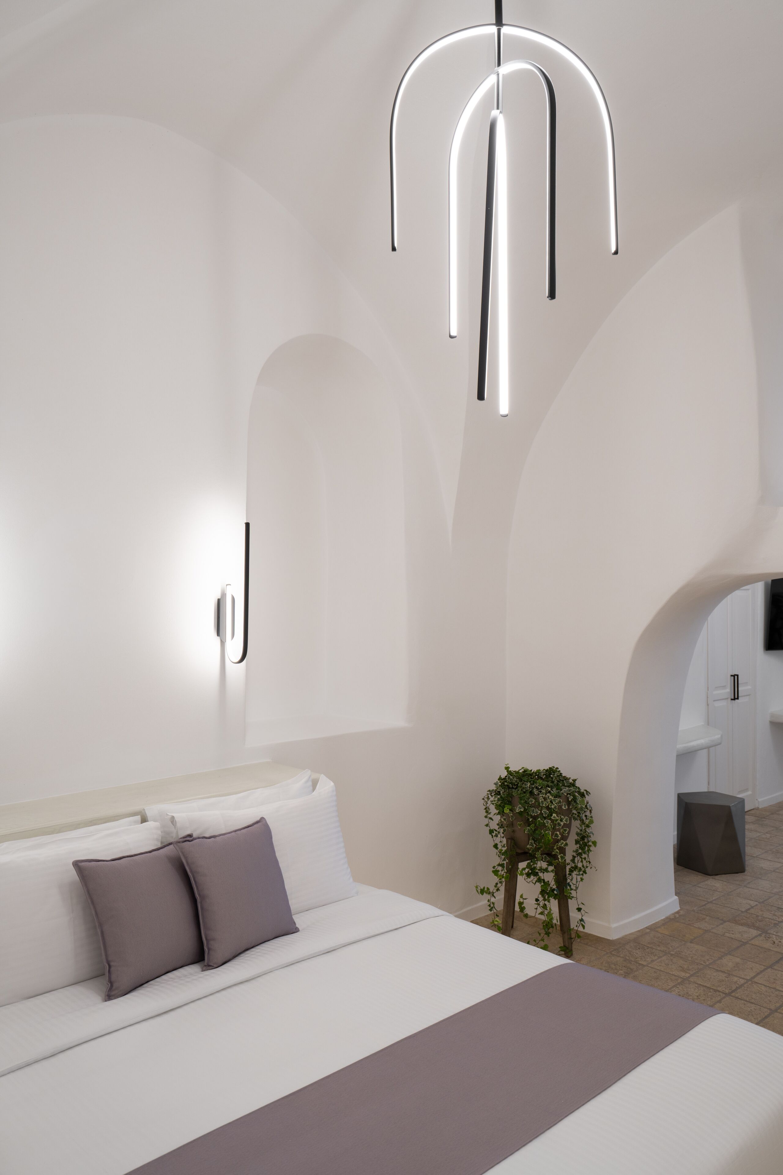A modern bedroom with a large bed, a bedside table, a hanging light fixture, and a small plant.