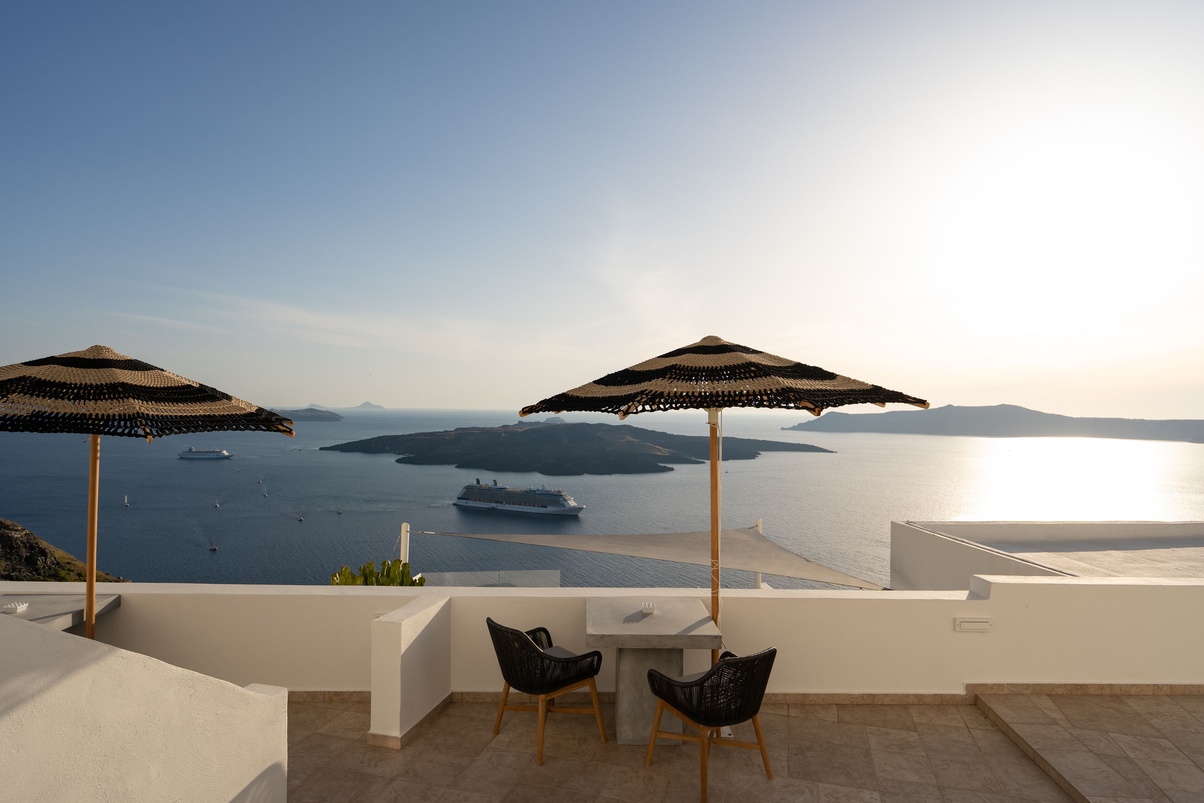 Una terraza con dos sombrillas, una mesa y sillas, con vistas a una masa de agua donde se ve un barco y varias islas.