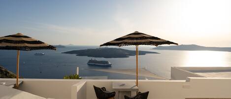 A terrace with two umbrellas, a table, and chairs overlooking a body of water with a ship and islands.