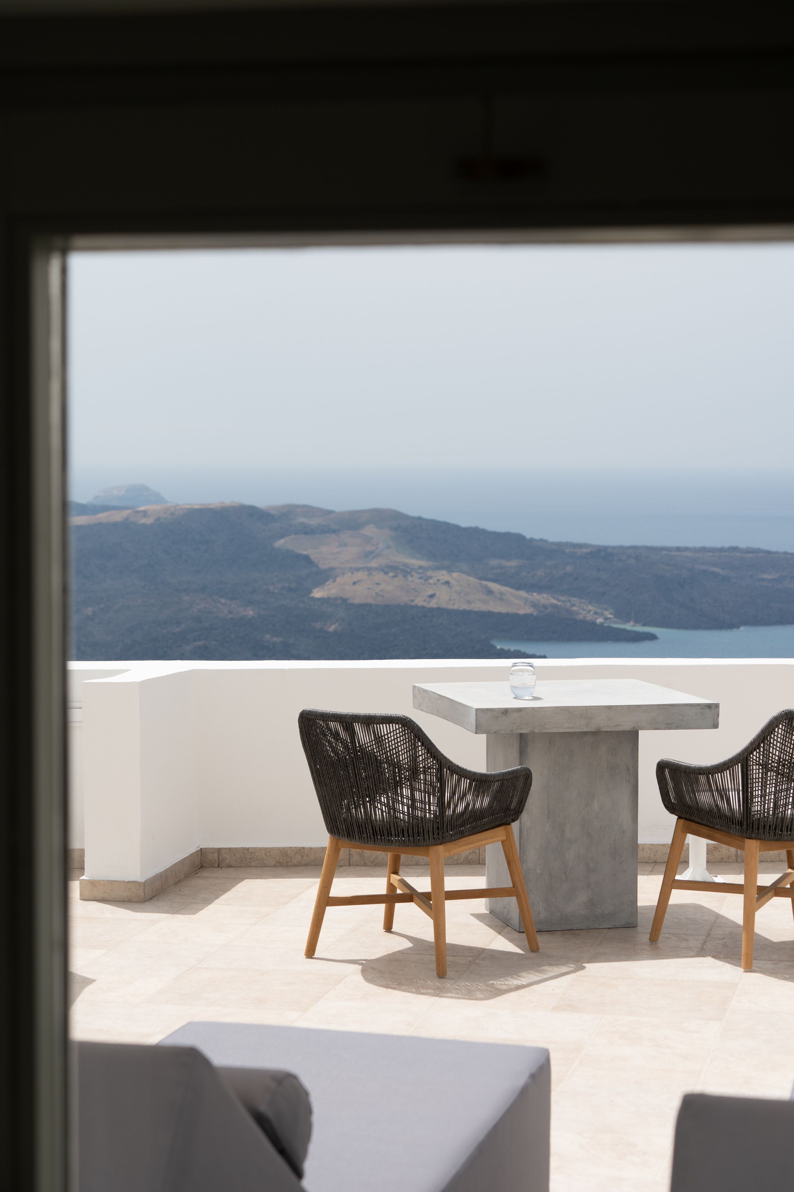 A balcony with two wicker chairs and a table, overlooking a scenic landscape with hills and a body of water.