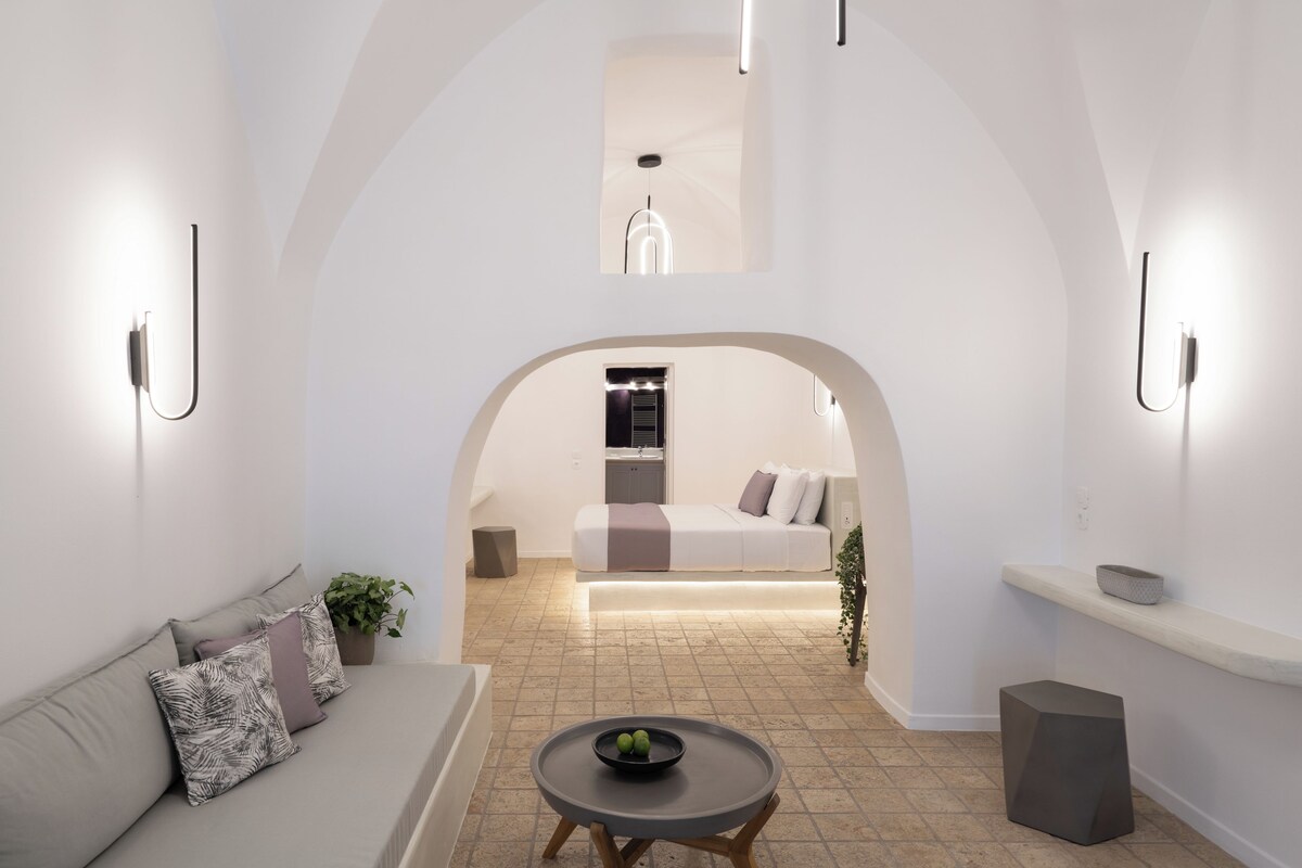 A modern interior with a sofa, a round table with a bowl, and a kitchen visible through an archway.