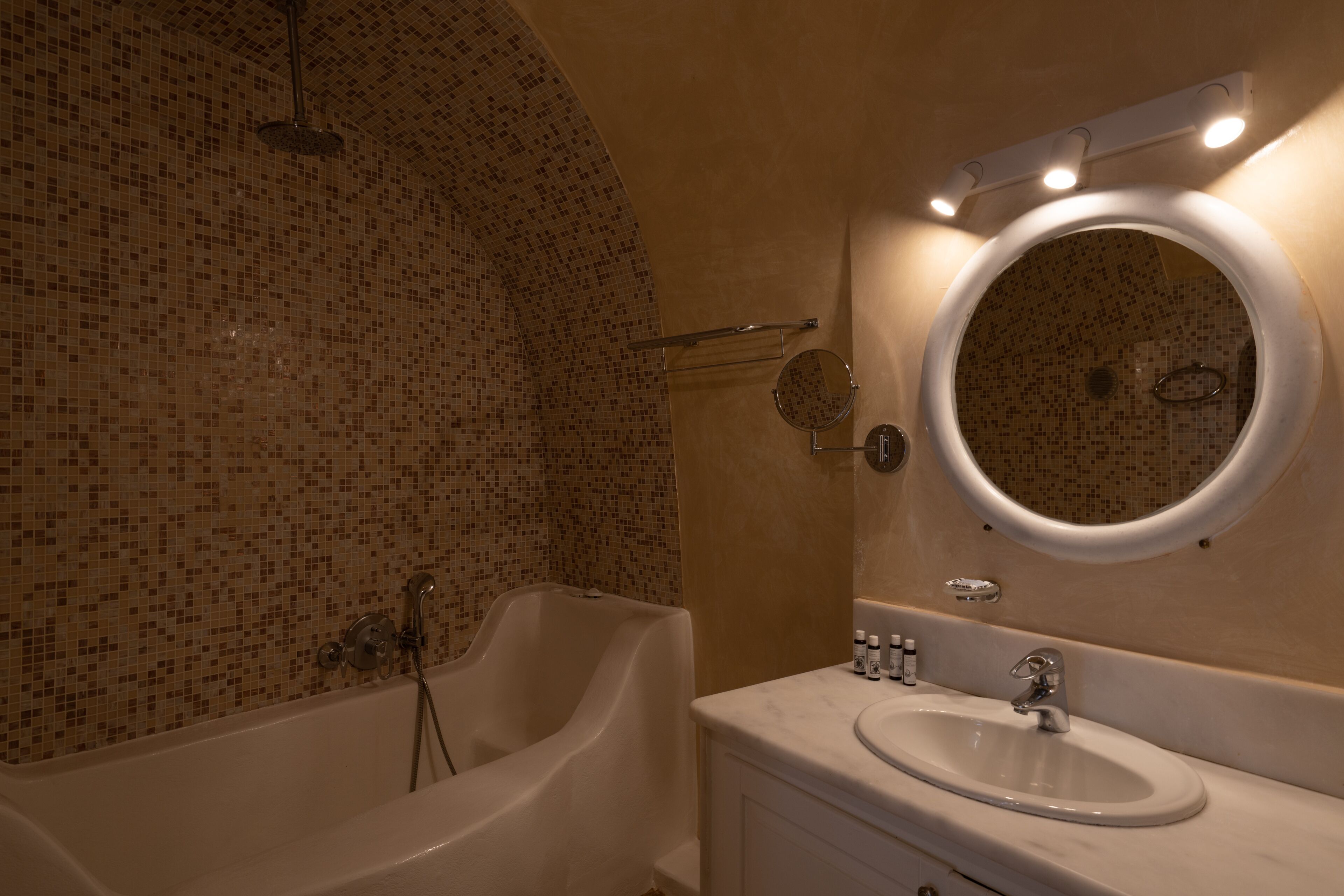 A bathroom with a mosaic-tiled wall, a freestanding bathtub, a round mirror with lights, and a sink with toiletries.