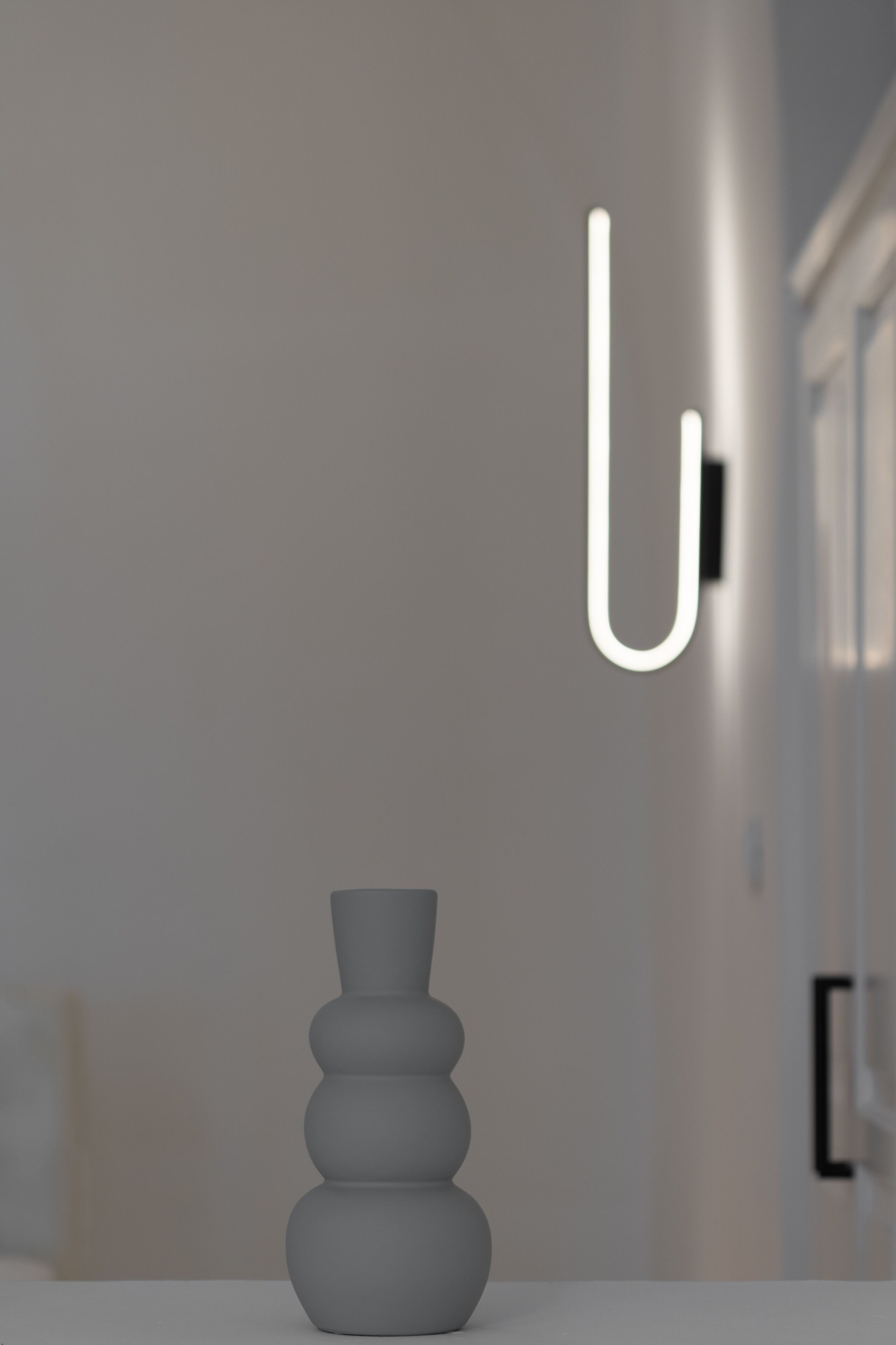 A modern interior with a minimalist design, featuring a white vase on a table and a simple, clean wall with a single light fixture.