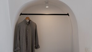 Una habitación con una pared blanca, un perchero de madera con una camisa gris y un dispositivo electrónico negro sobre una estantería.