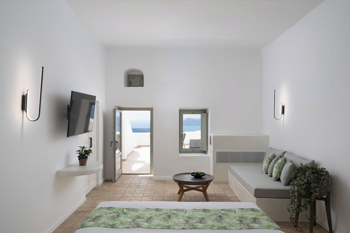 Ein modernes, minimalistisches Zimmer mit einem Bett, einem kleinen Tisch, einer Couch und einem an der Wand befestigten Fernseher. Zwei Fenster bieten Ausblicke ins Freie.