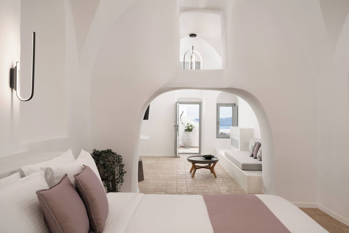 Ein modernes Schlafzimmer mit einem großen Bett, einem kleinen runden Tisch und einem Blick durch einen Bogen auf einen Balkon mit Sitzgelegenheit.