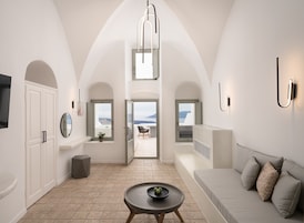 A modern interior with a high ceiling, arched doorways, a seating area with a sofa and cushions, a small table with a bowl, and a view through the windows.