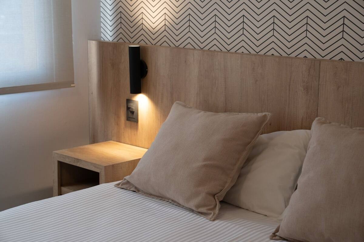 A neatly made bed with two pillows and a wooden bedside table with a wall-mounted light.