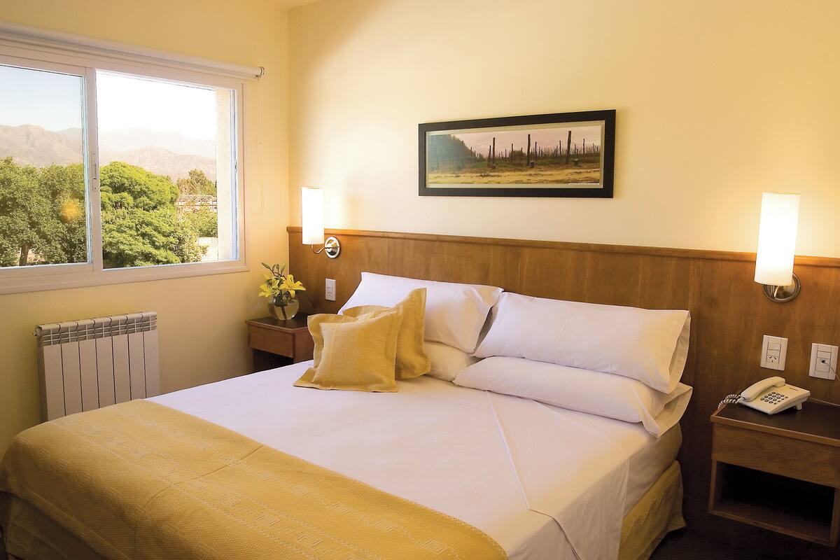 A hotel room with a bed, a nightstand with a phone, a framed picture on the wall, and a window with a view of trees and mountains.