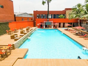 Piscina con sillas de descanso y sombrillas, rodeada de edificios con paredes de color rojo y naranja.