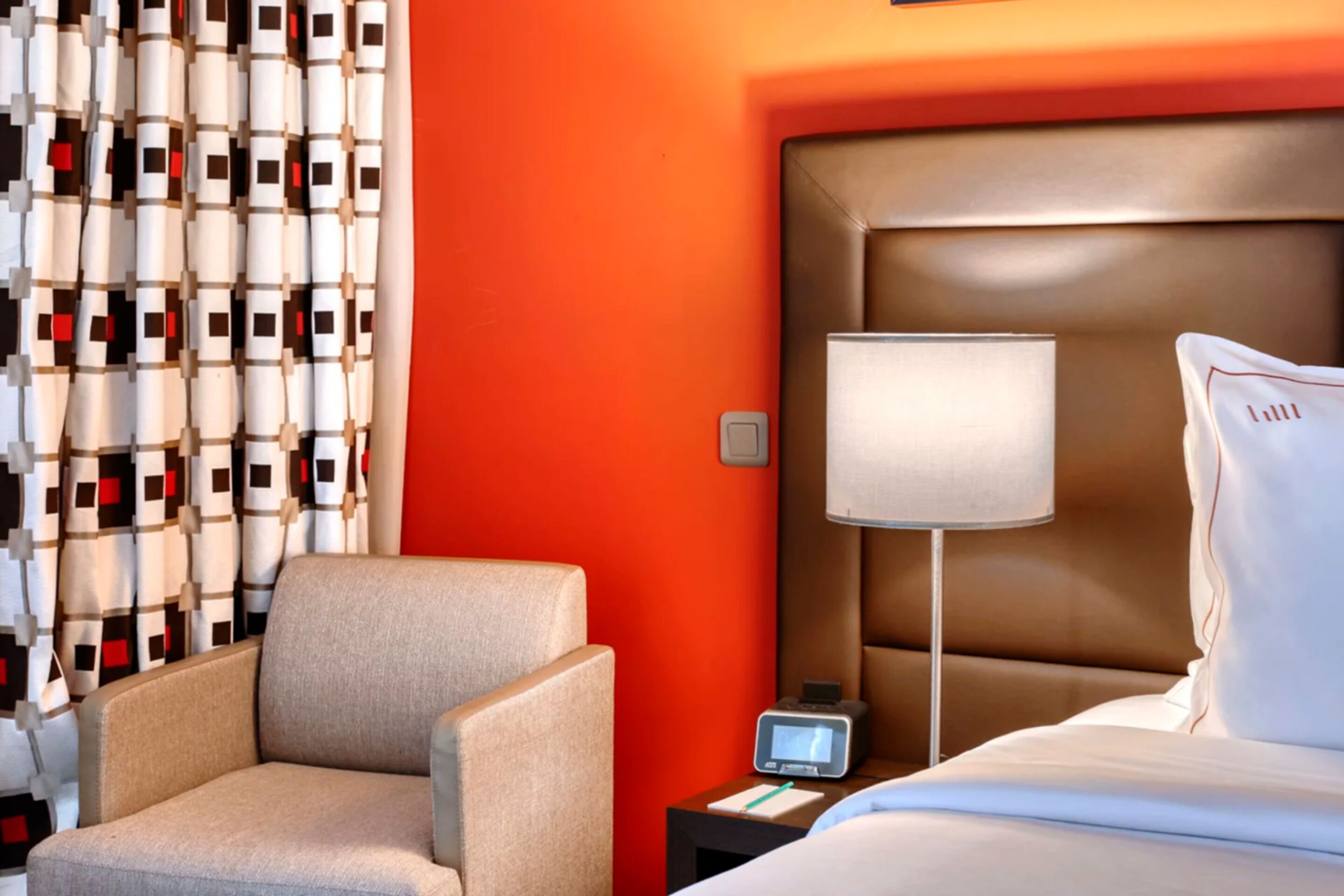 Habitación de hotel con una cama grande, una mesita de noche con lámpara, una silla y una pared con cortina estampada.