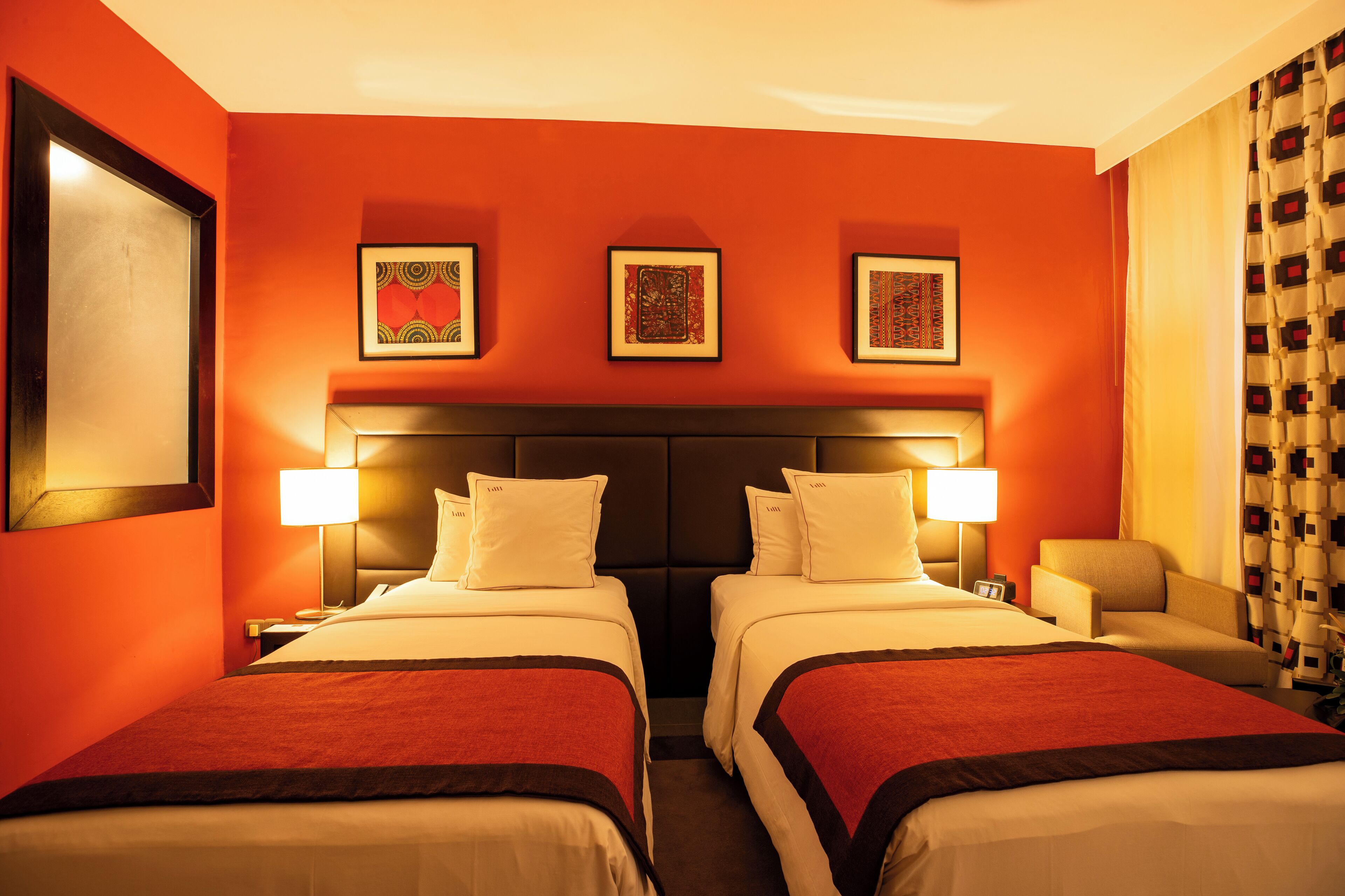 A hotel room with two beds, a headboard, bedside lamps, and three framed artworks on the wall.