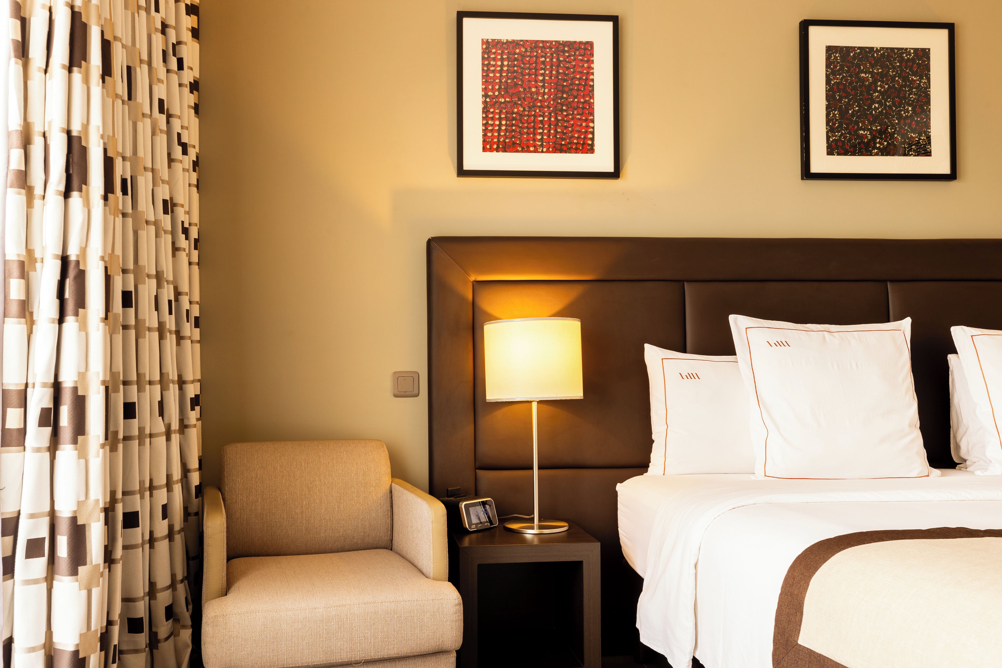 A hotel room with a bed, a chair, a nightstand with a lamp, and two framed pictures on the wall.