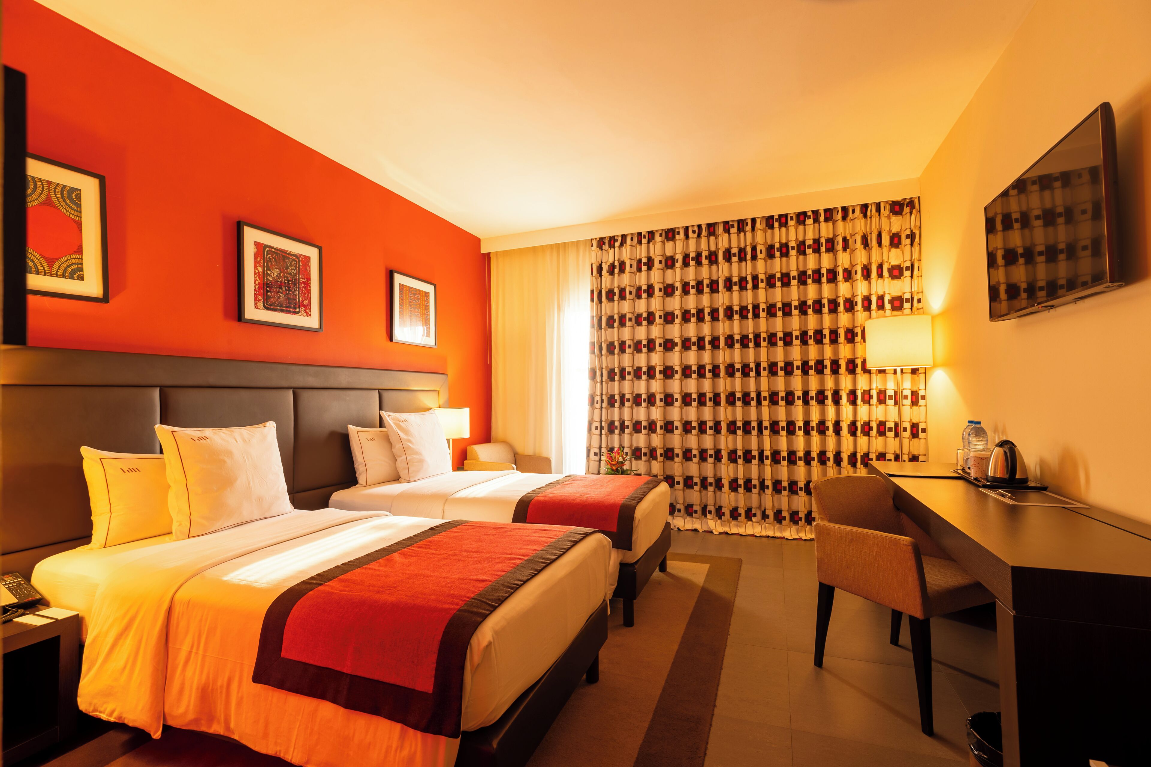Ein Hotelzimmer mit zwei Betten, einem Schreibtisch, einem Stuhl und einem großen Fenster mit gemusterten Vorhängen.
