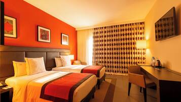 Ein Hotelzimmer mit zwei Betten, einem Schreibtisch, einem Stuhl und einem großen Fenster mit gemusterten Vorhängen.