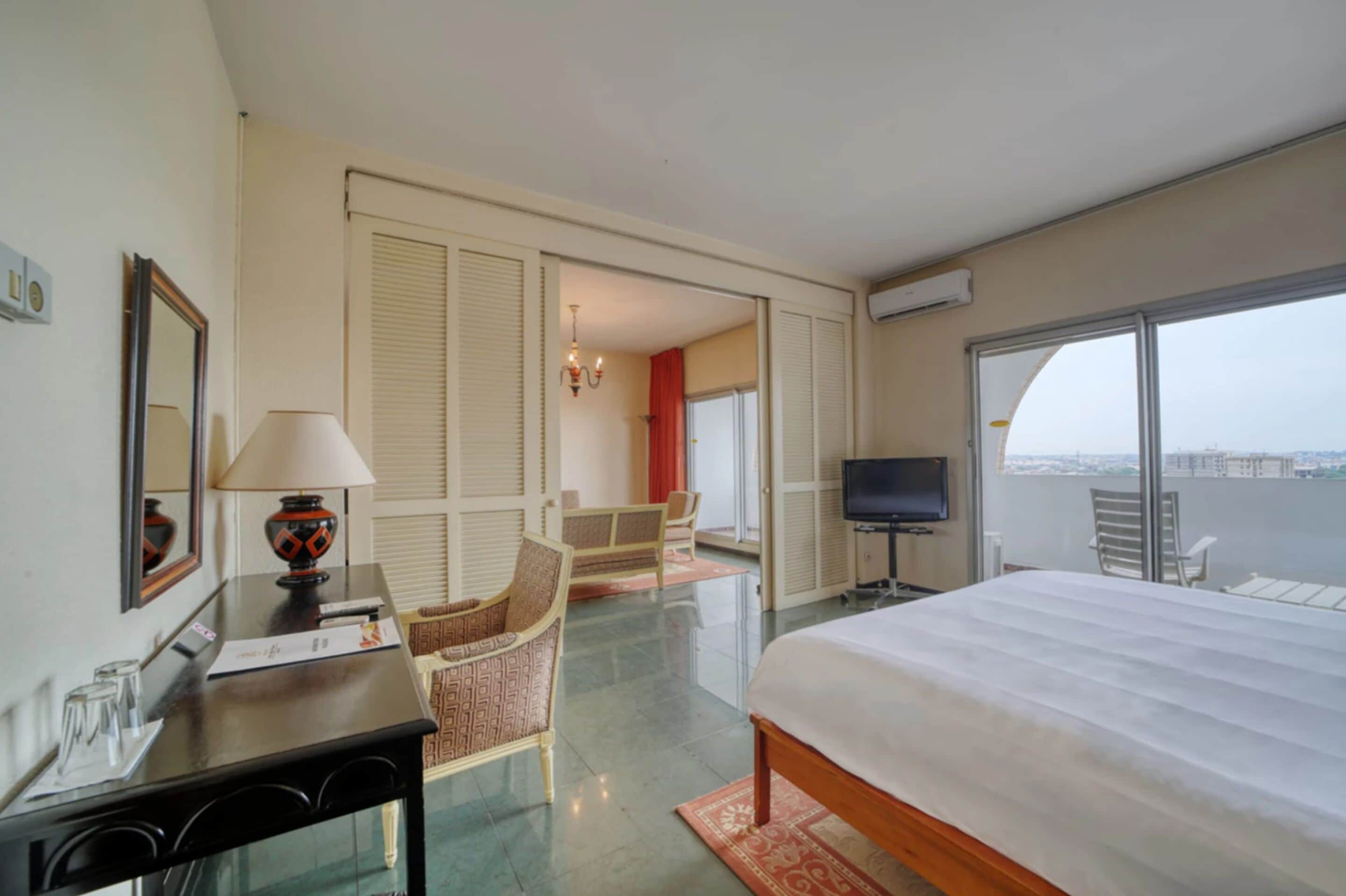 A hotel room with a bed, a desk with a lamp, a TV, a chair, and a view of the city through large windows.