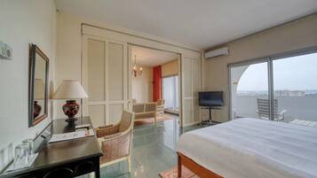 A hotel room with a bed, a desk with a lamp, a TV, a chair, and a view of the city through large windows.