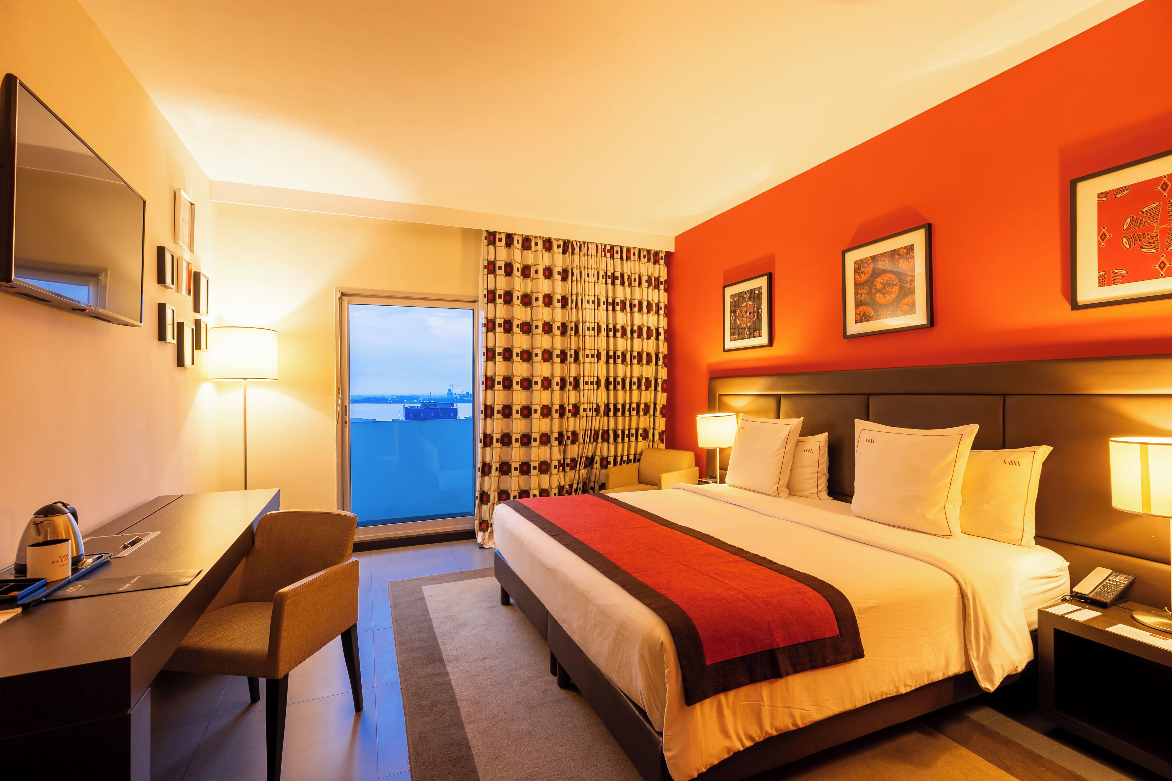 A hotel room with a bed, desk, chair, and a view of the city through the window.