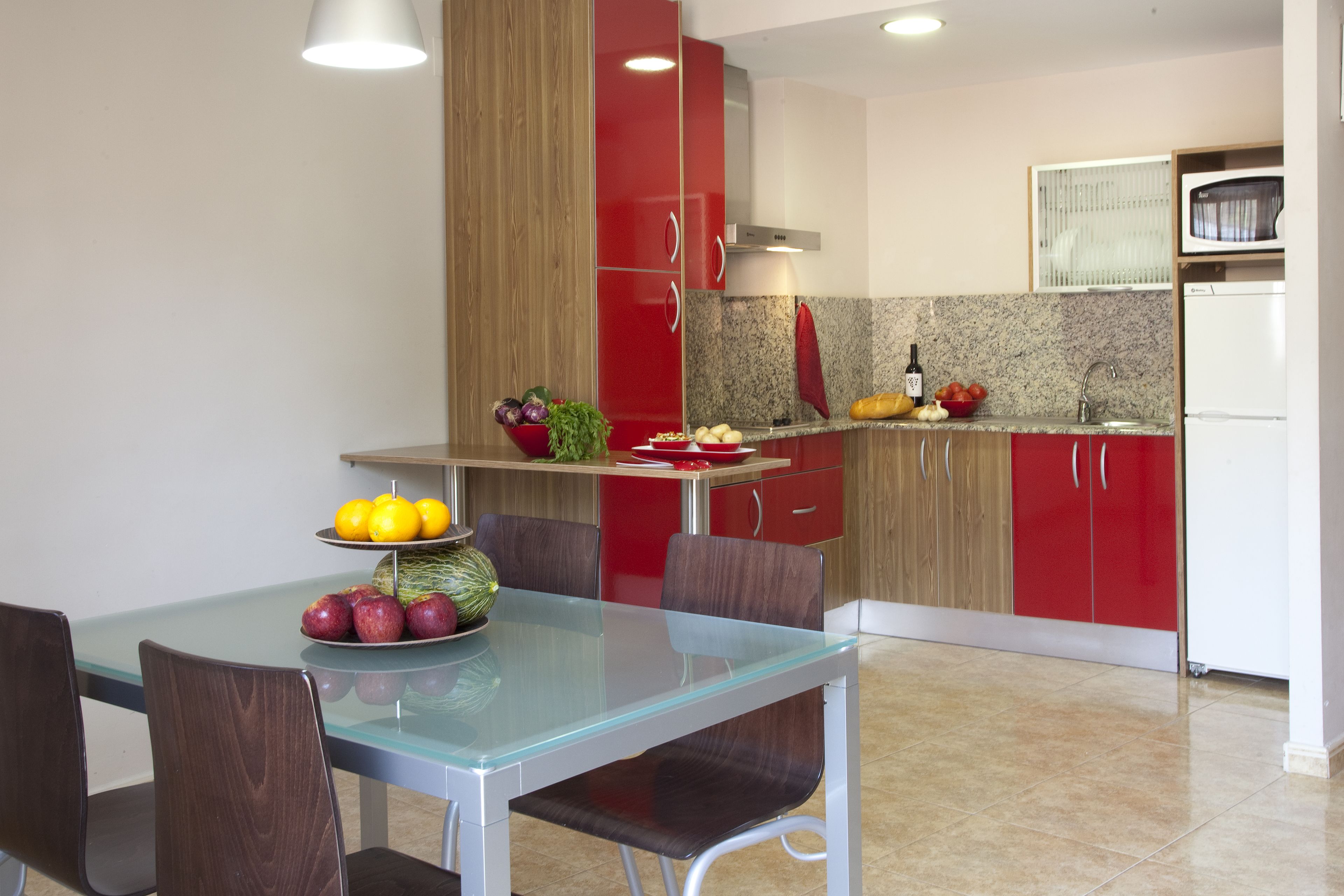 A modern kitchen with red cabinets, a glass dining table, and a variety of fruits and vegetables on the counter.