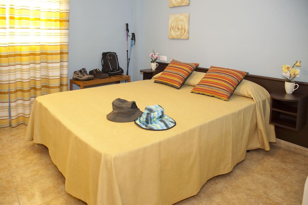 A neatly made bed with a yellow bedspread, two pillows, and a pair of hats on the bed. There is a small table with a lamp and a vase with flowers beside the bed.