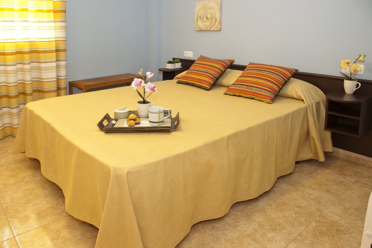 A neatly made bed with a yellow bedspread, a tray with a vase of flowers, and a bedside table with a plant and a cup.