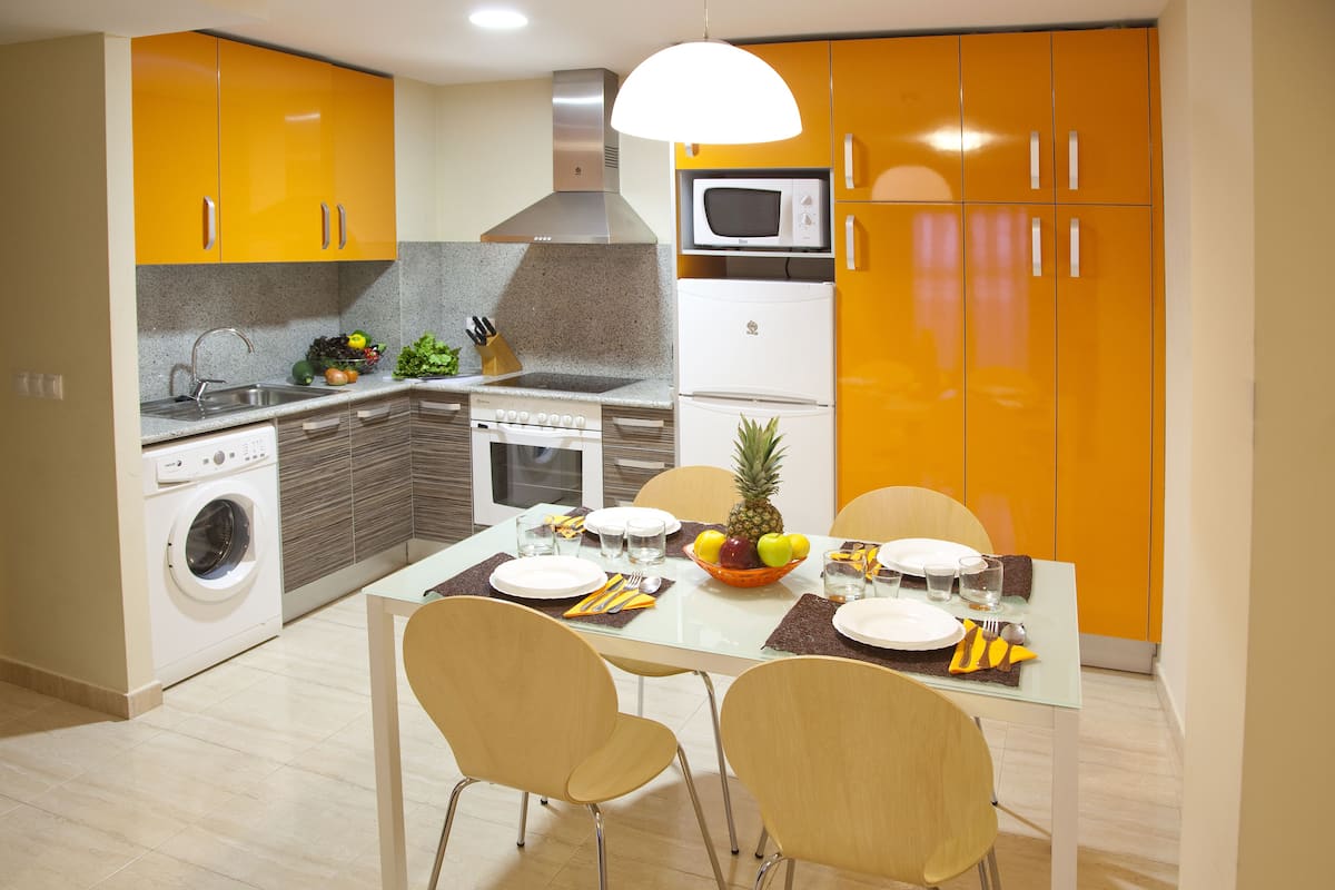 A modern kitchen with a dining area, featuring a washing machine, microwave, and a fruit bowl on the table.