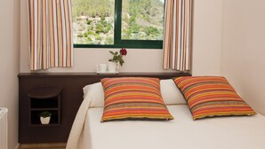 Een slaapkamer met een bed, twee kussens, een raam met gordijnen, een klein plankje met een plant en een vaas met een bloem.