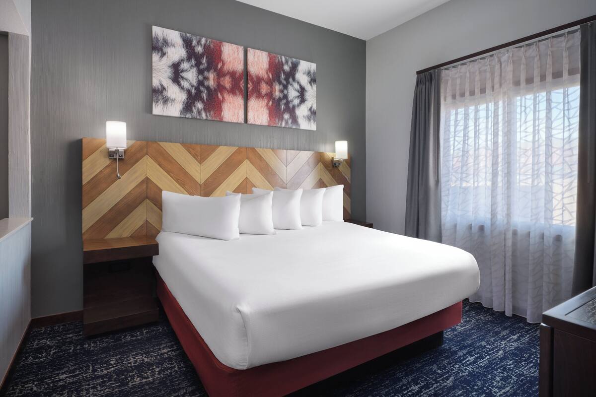A hotel room with a large bed, a wooden headboard, two wall-mounted lights, and a window with sheer curtains.